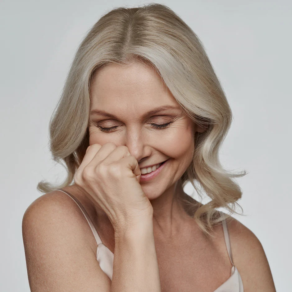 Mujer de edad avanzada sonriendo cabizbaja mientras se tapa parte de la sonrisa con la mano derecho 
